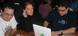 Derek Powazek, Heather Champ, and Tantek elik are seated at a table.  Derek is looking off to the left with an expression of diabolical amusement; Heather is speaking to someone outside the frame, her right hand to her cheek; and Tantek types away on his new Macintosh iBook.